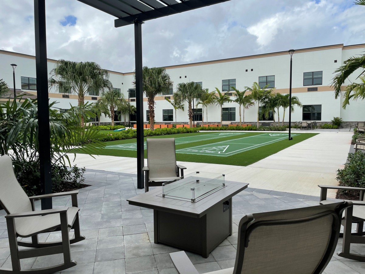 A modern patio area with a fire pit table and outdoor seating overlooking a landscaped lawn with palm trees.