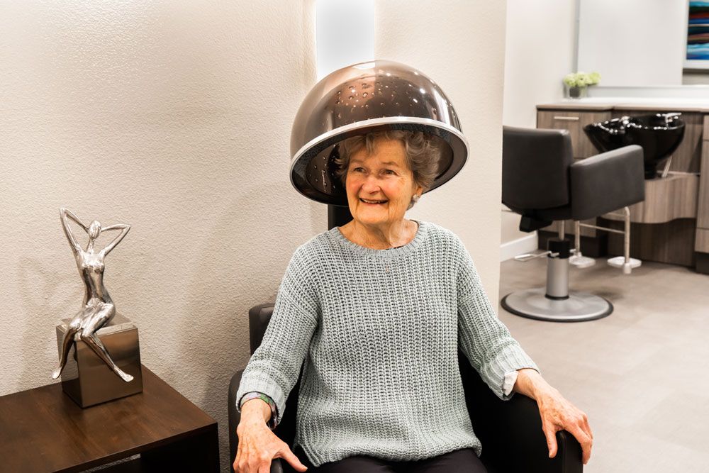 An elderly woman smiles while sitting under a hair dryer at a salon.