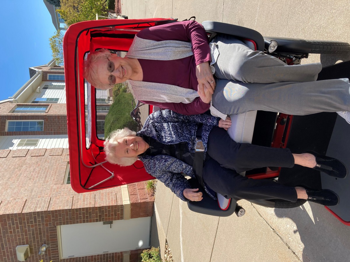 Two elderly women seated on a red tricycle outdoors on a sunny day.