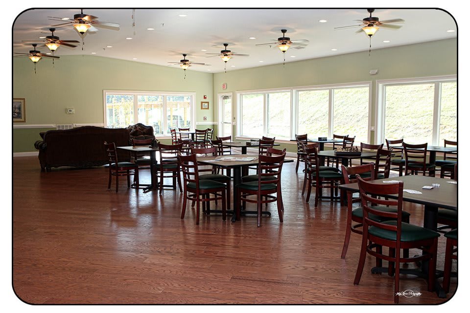An empty dining area with wooden floors and tables surrounded by chairs, illuminated by natural light.