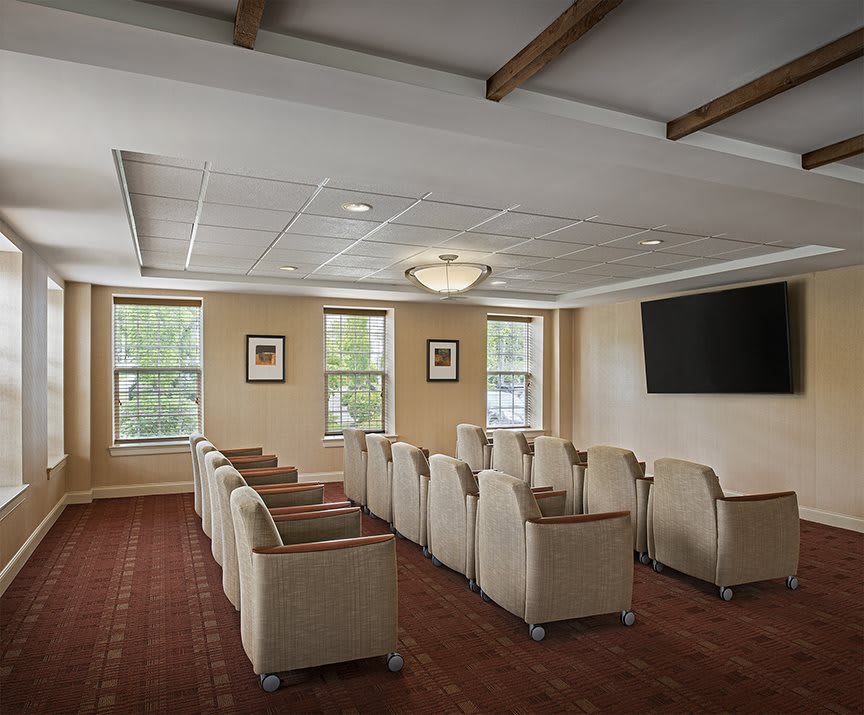 A meeting room with rows of chairs facing a large television, featuring carpeted flooring and windows letting in natural light.
