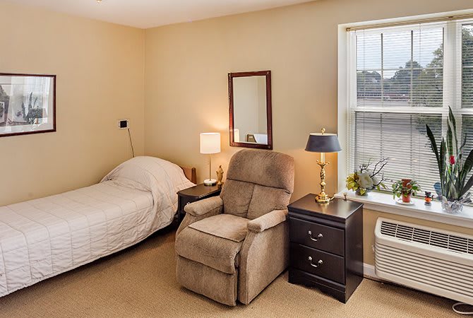 A cozy bedroom featuring a small bed, comfortable armchair, and a nightstand with a lamp by the window.