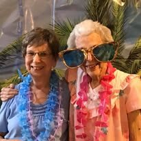 Two elderly women enjoying a Hawaiian-themed party with leis and oversized sunglasses.