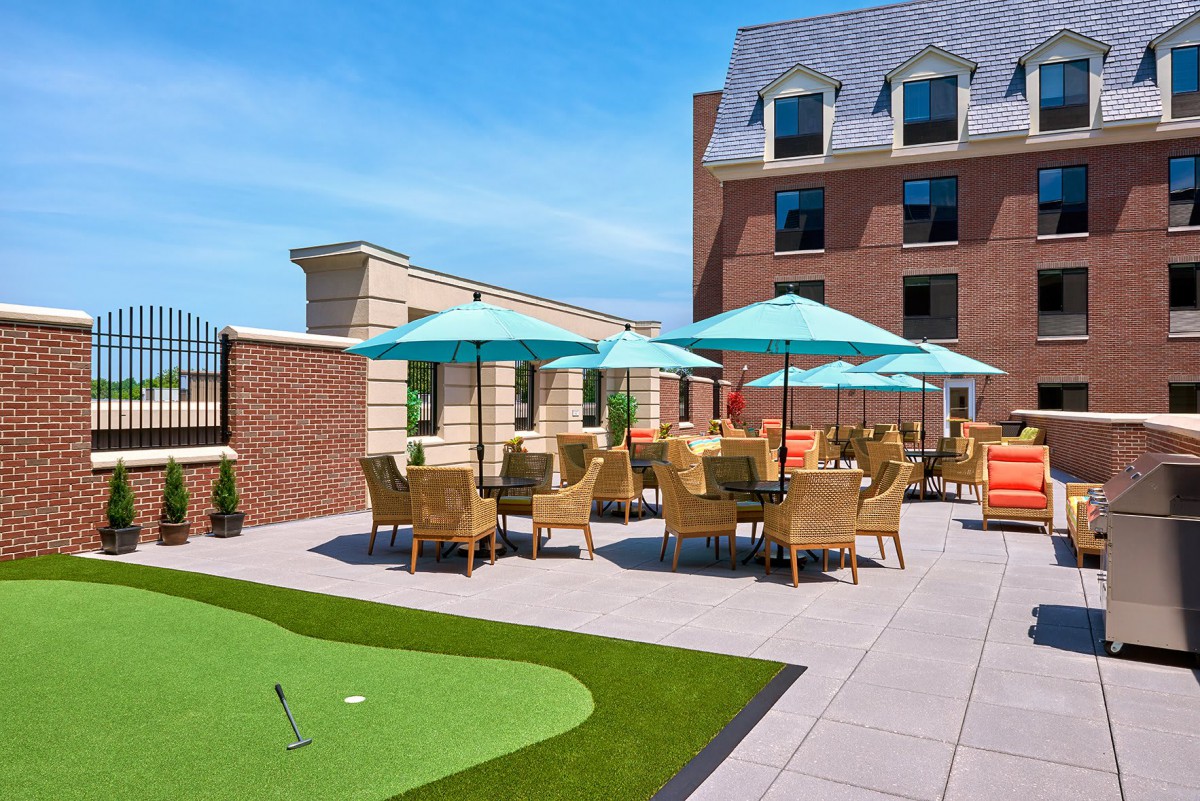 A rooftop patio with wicker furniture and umbrellas under a clear blue sky, adjacent to a brick building.
