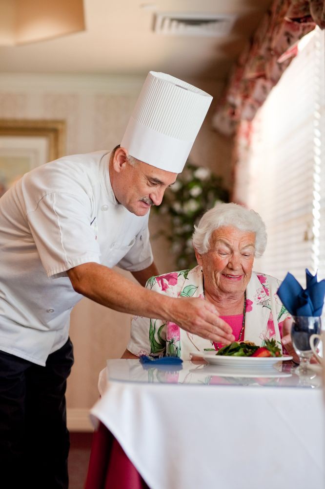 A chef in a white uniform serves a meal to an elderly woman sitting at a restaurant table.