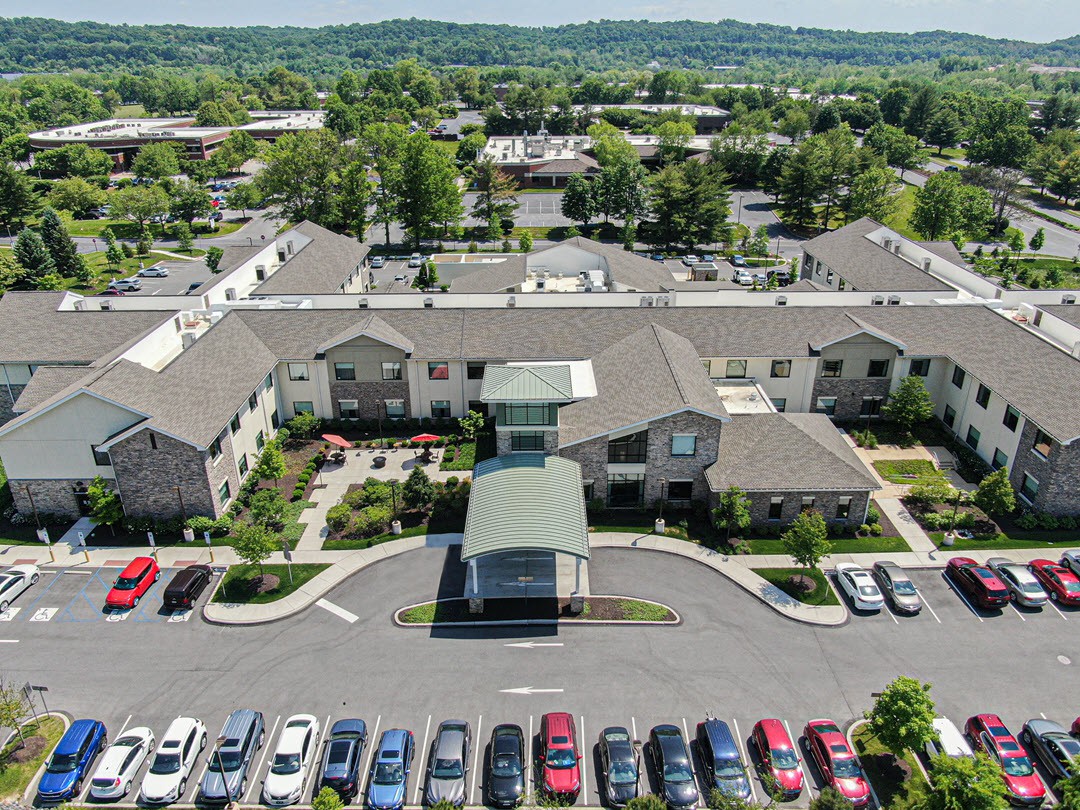 An aerial view of a complex with multiple buildings and a parking lot full of cars, surrounded by greenery.