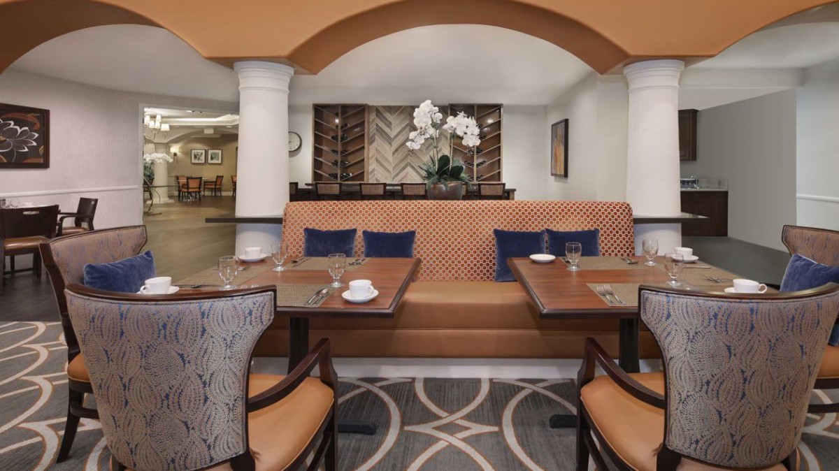 A cozy restaurant interior featuring banquette seating with decorative pillows and elegant table settings, complemented by a centerpiece of orchid flowers.