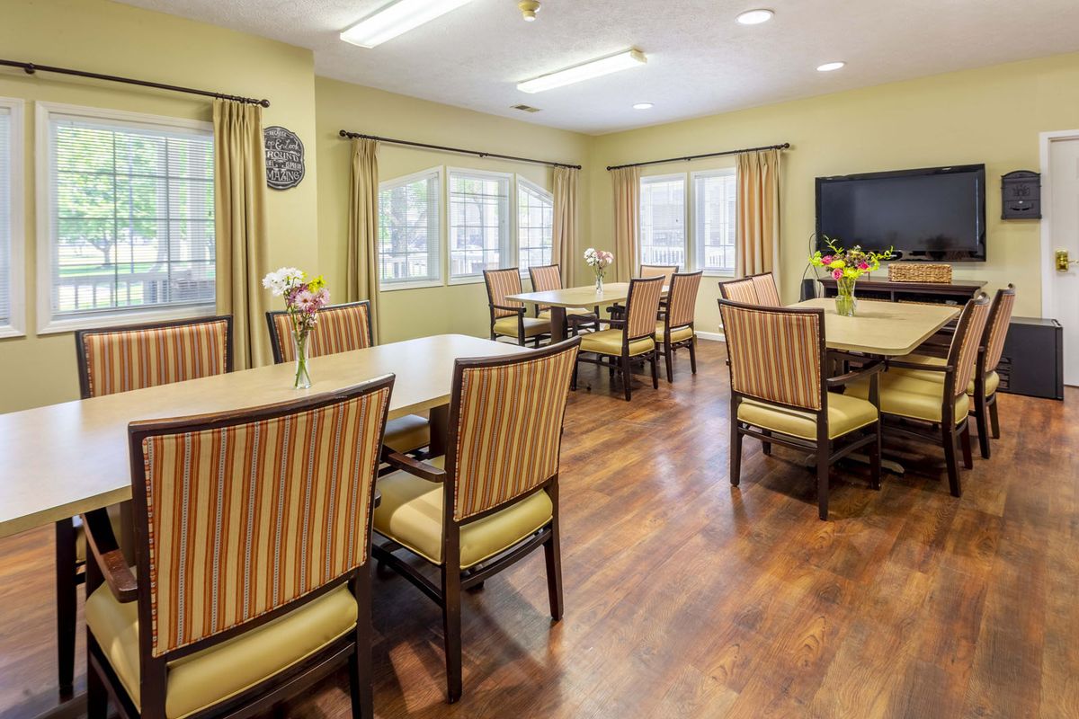 A well-lit dining room with multiple tables and chairs, featuring large windows and wooden flooring.