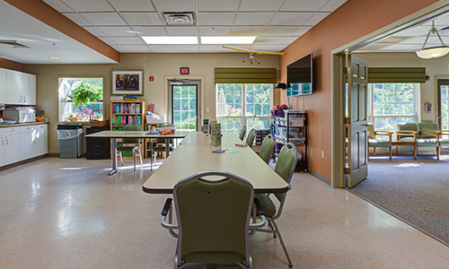 A community room with tables and chairs, featuring a television, bookshelves, and large windows.