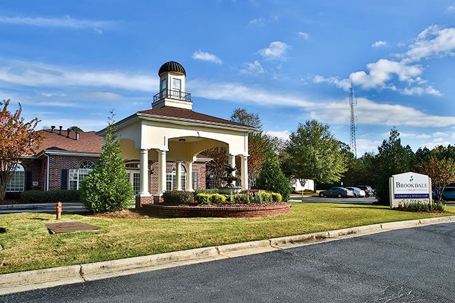 A modern building featuring a clock tower, surrounded by a landscaped area and a parking lot under a blue sky.