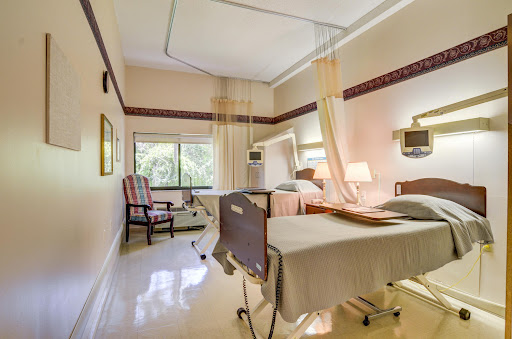 A well-designed hospital patient room featuring two beds, a sitting area, and natural light from the window.