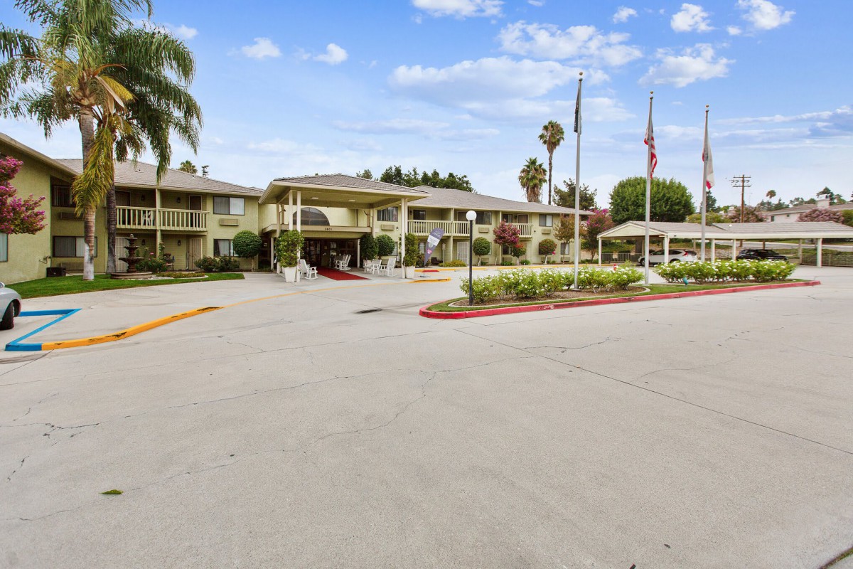 The entrance of a hotel with a parking area, surrounded by palm trees and flags.