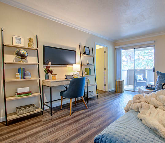 A modern living space featuring a home office setup with a desk and chair, complemented by a stylish bookshelf and natural light from the balcony.