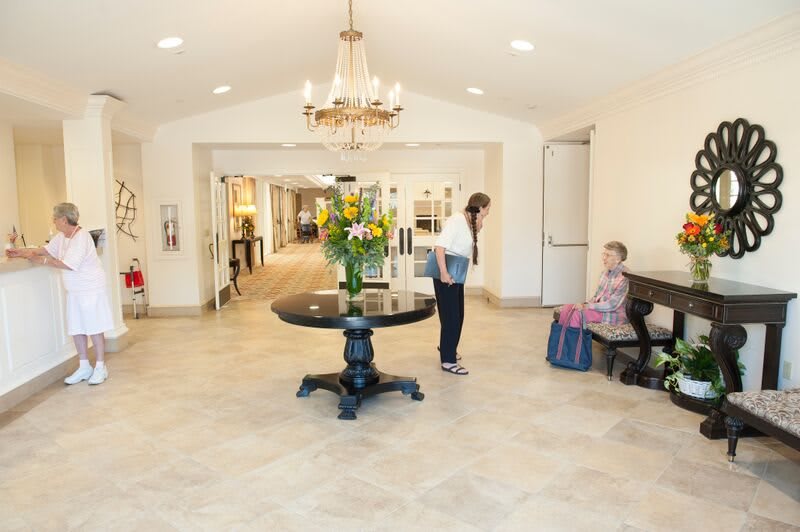 A spacious lobby with a chandelier, flowers on a round table, and two people interacting in the reception area.