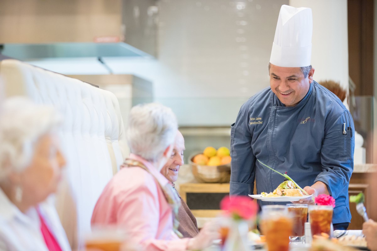 A chef serving a meal to elderly patrons in a restaurant setting.