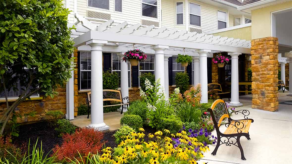 A beautifully landscaped garden with colorful flowers, benches, and a white pergola supported by columns in front of a building.