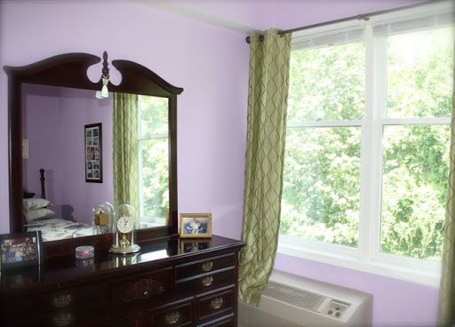 A cozy bedroom featuring a dark wooden dresser, a large mirror, and flowing green curtains by the window.