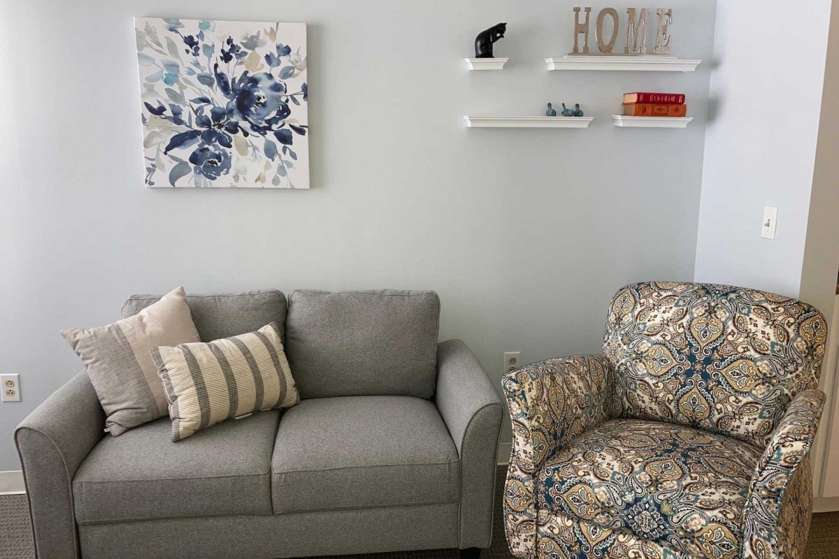 A cozy living room with a gray sofa, patterned armchair, wall art, and decorative shelves.