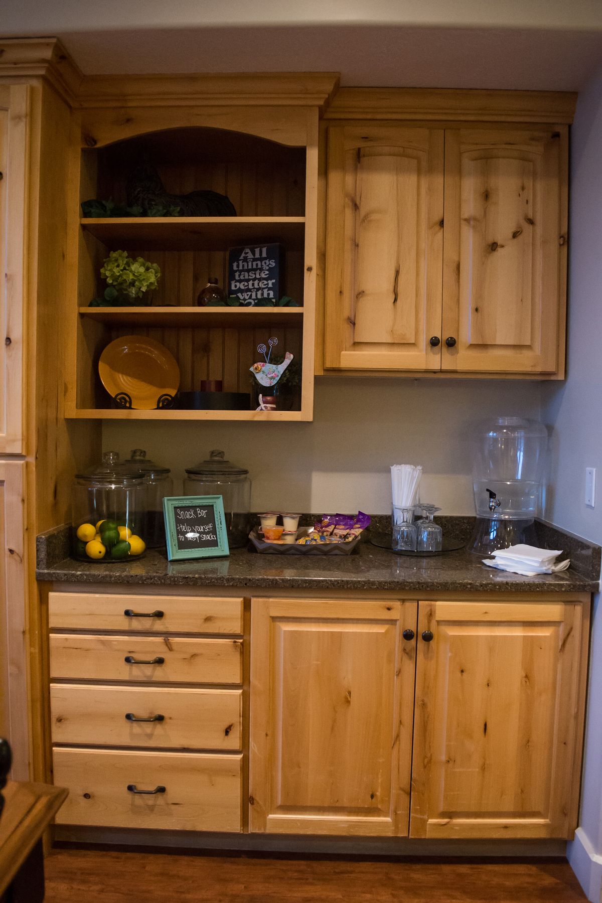 A kitchen area featuring wooden cabinets with open shelves, a countertop with fruit and a drinks dispenser.