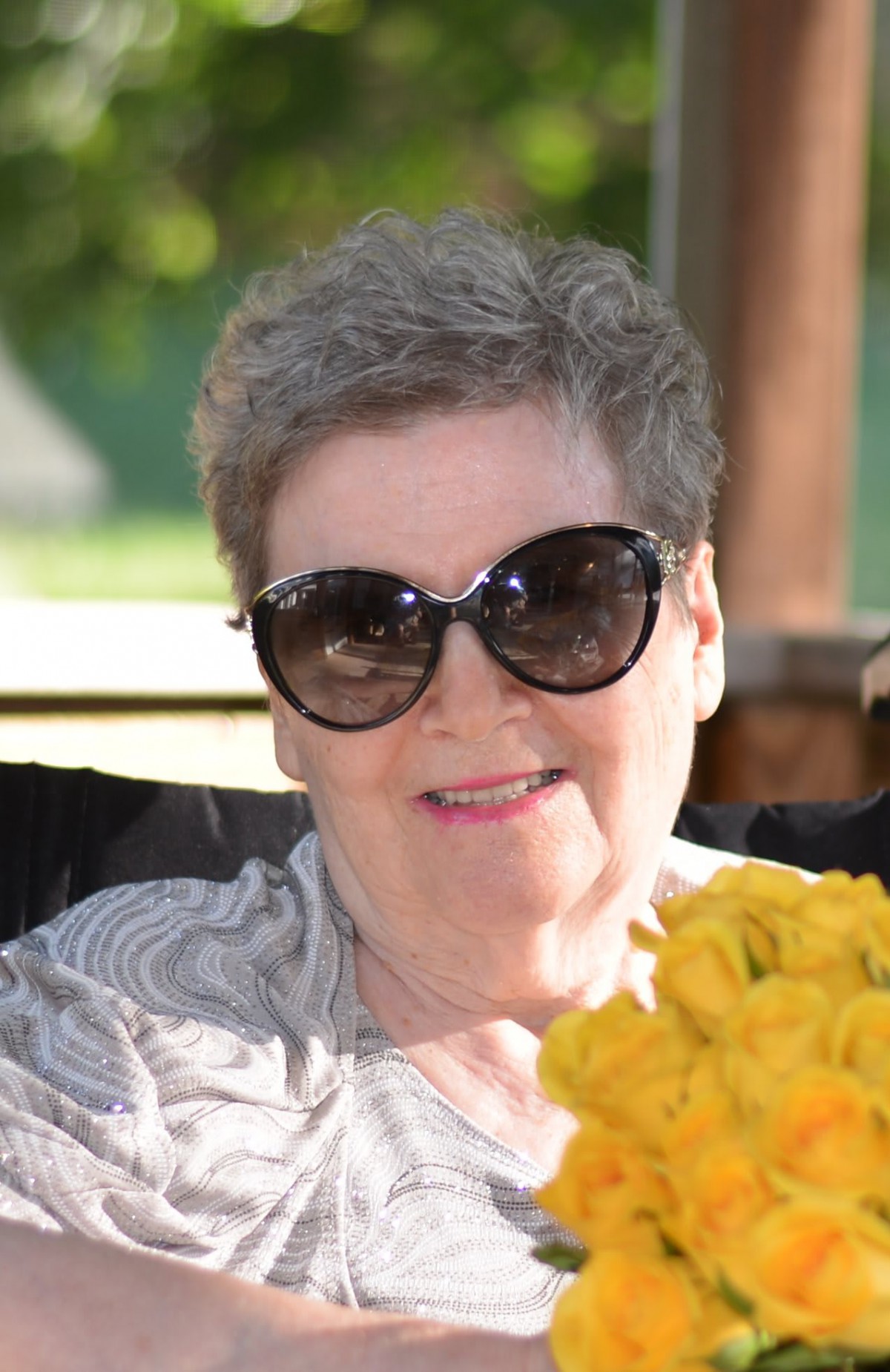 An older woman wearing sunglasses, smiling while sitting outside with flowers in front of her.