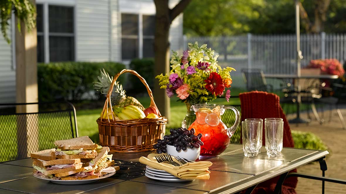 A refreshing summer picnic setup with a basket of fruits, a pitcher of juice, and fresh flowers on a table outdoors.