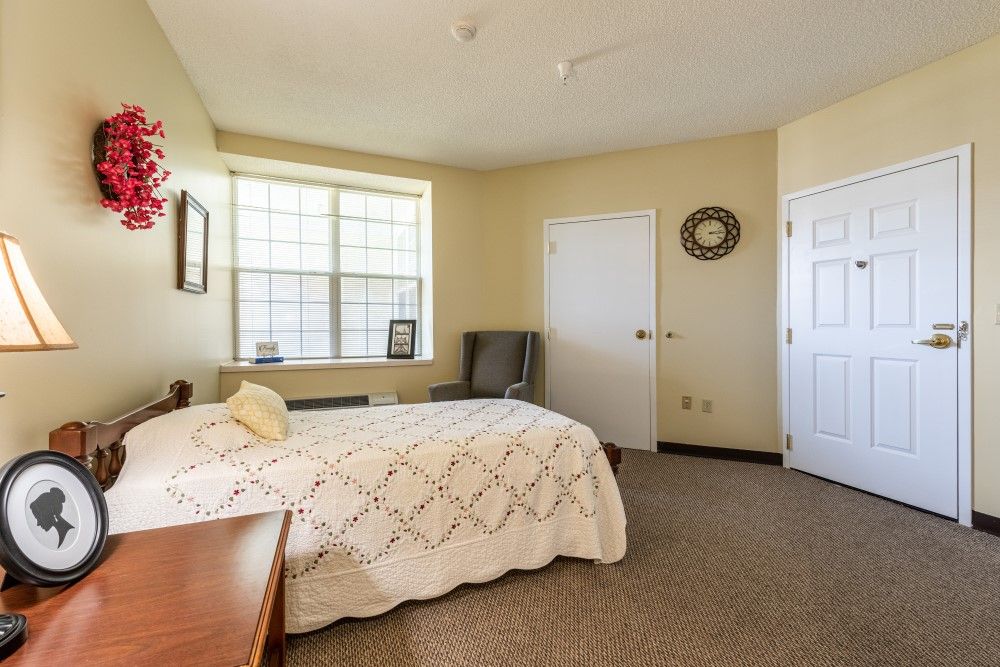 A cozy bedroom interior with a neatly made bed, armchair, and a window allowing natural light.