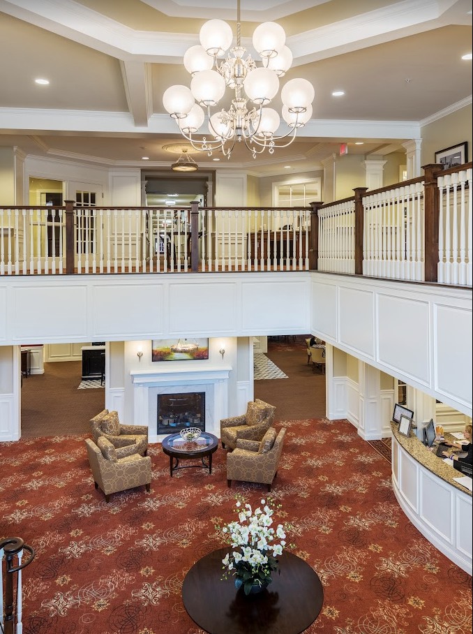A spacious and elegantly designed lobby featuring a chandelier, armchairs, and a fireplace with a balcony overlooking the area.