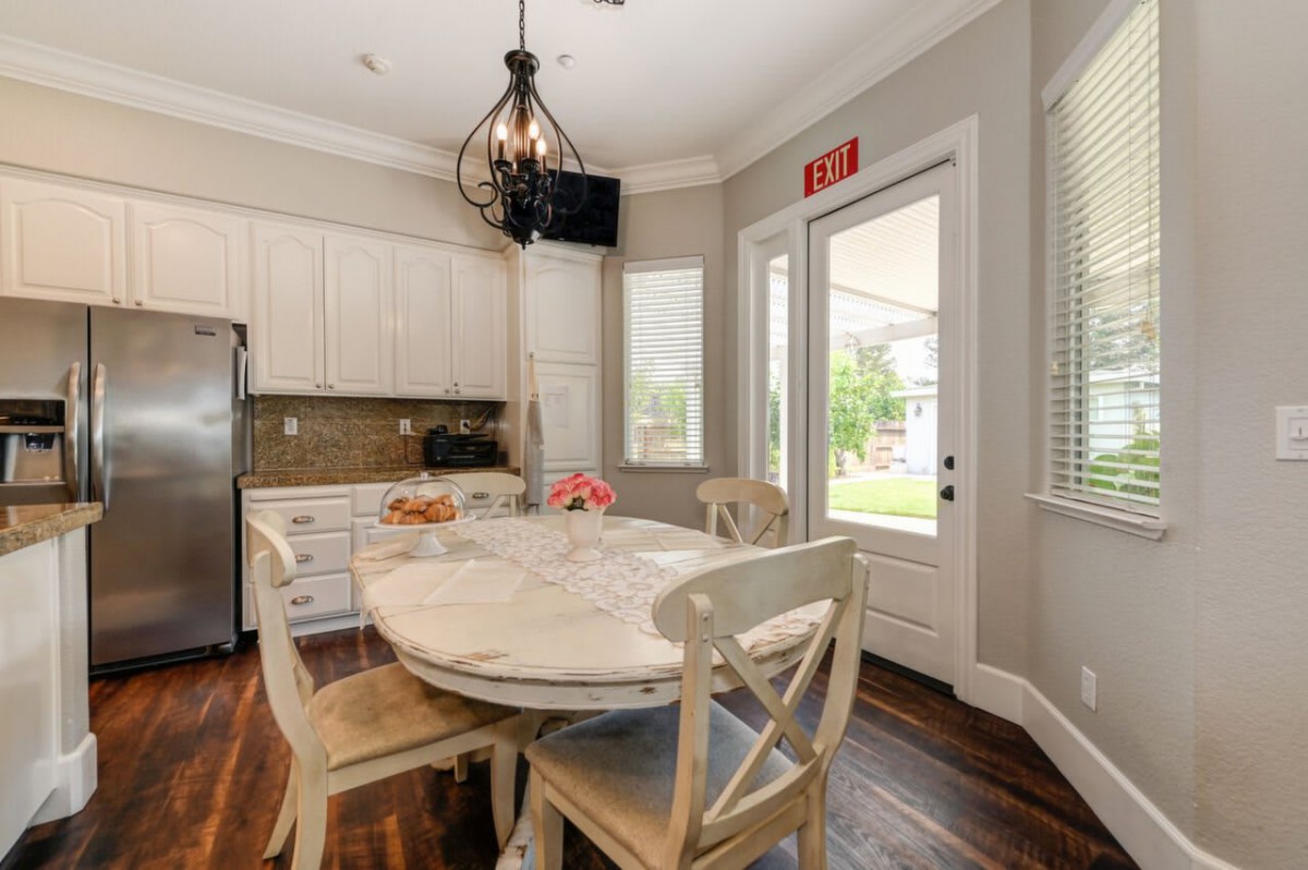 A cozy kitchen with a round dining table and chandelier, featuring white cabinets and hardwood floors bathed in natural light.