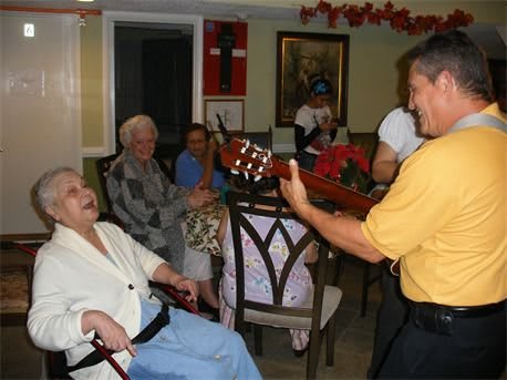 A person plays guitar and sings to a group of seniors indoors.