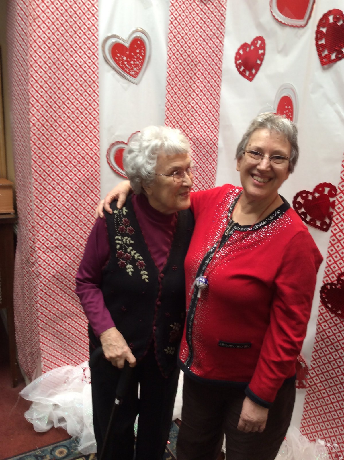 Two women smiling and hugging in front of a heart-themed backdrop.