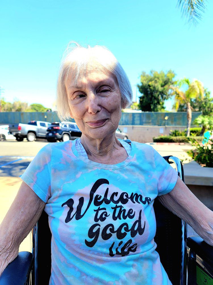 An elderly woman sitting outdoors on a sunny day, wearing a t-shirt with the phrase 'Welcome to the good life.'