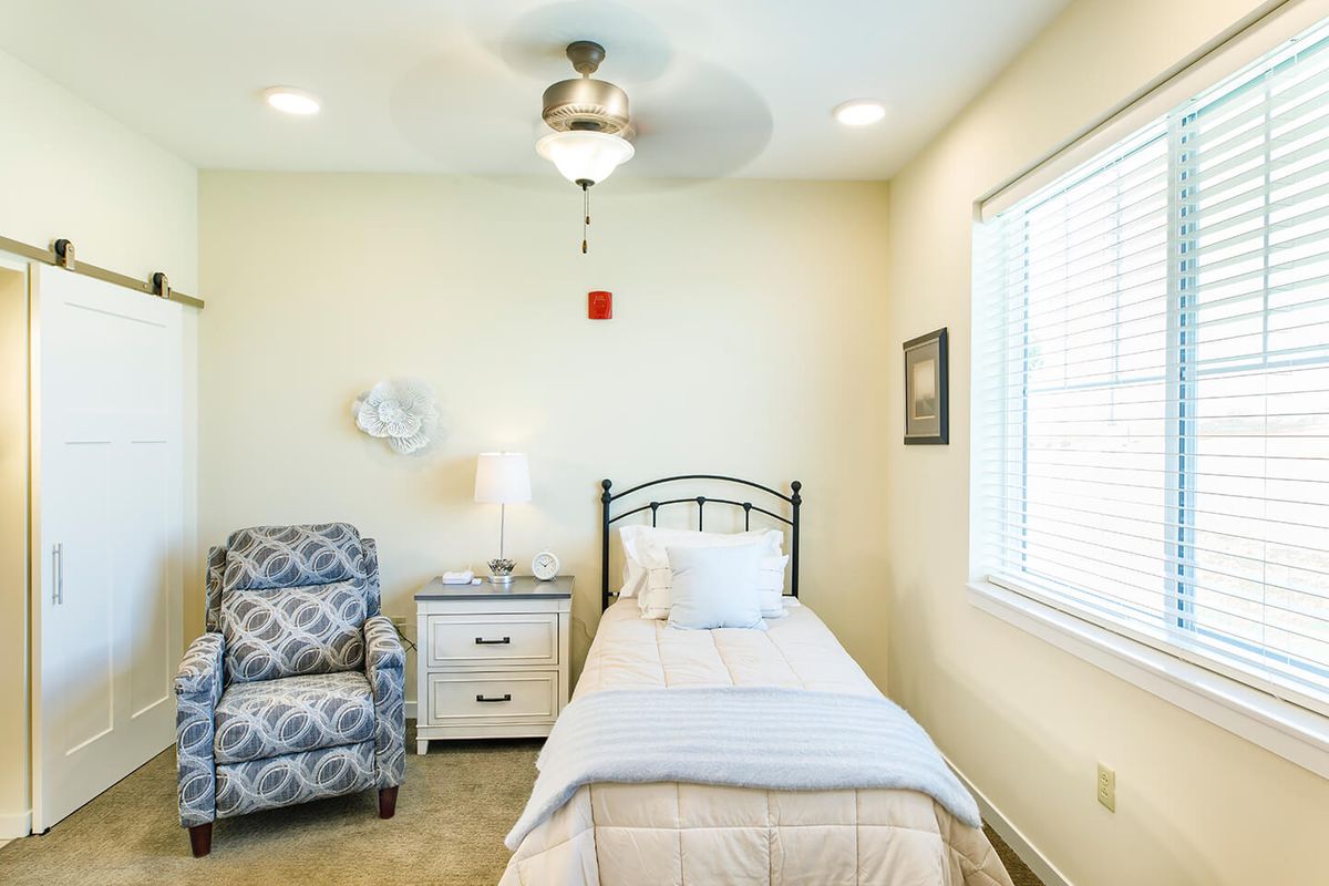 A minimalist bedroom with a single bed, armchair, and nightstand under a ceiling fan.