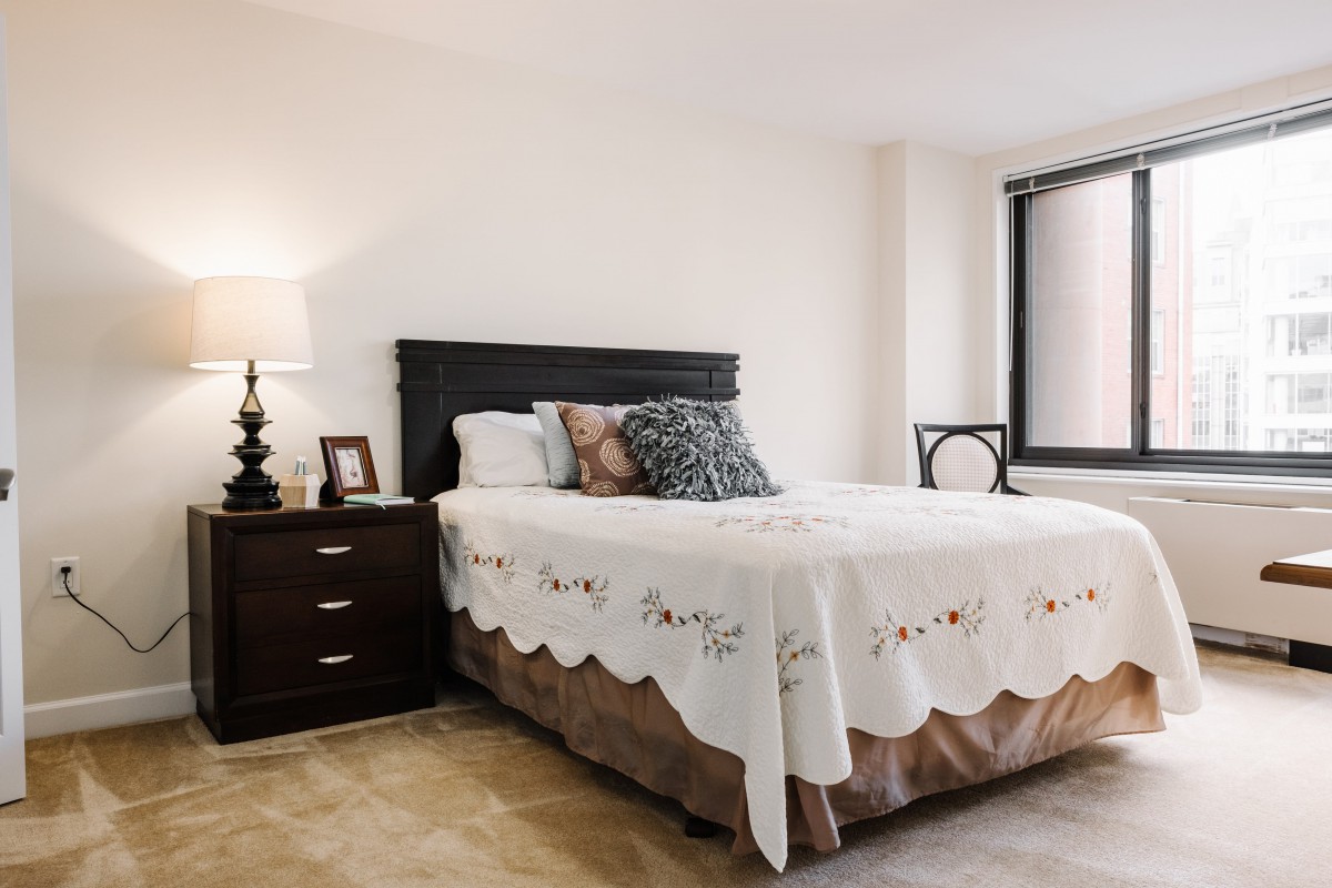 A cozy bedroom featuring a neatly made bed with decorative pillows, a nightstand with a lamp, and ample natural light from a large window.