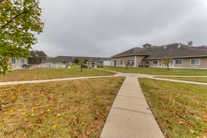 A pathway leads through a grassy area towards residential buildings under an overcast sky, with autumn leaves scattered on the ground.