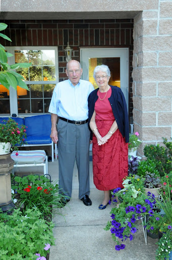 An elderly couple stands together in a garden, surrounded by colorful flowers.