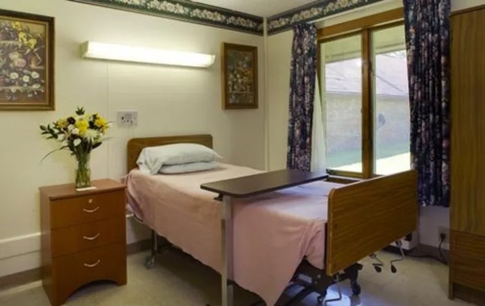 A hospital room with a neatly made patient bed and a small bedside table under a window.