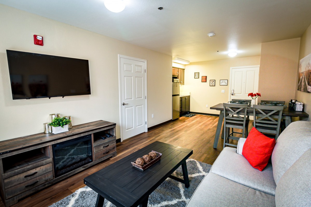 A modern living room with a cozy sofa, coffee table, and wall-mounted television, leading to an open dining area.