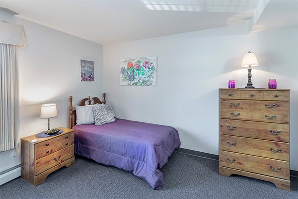 A cozy bedroom with purple bedding, a wooden dresser, and a nightstand adorned with decor.
