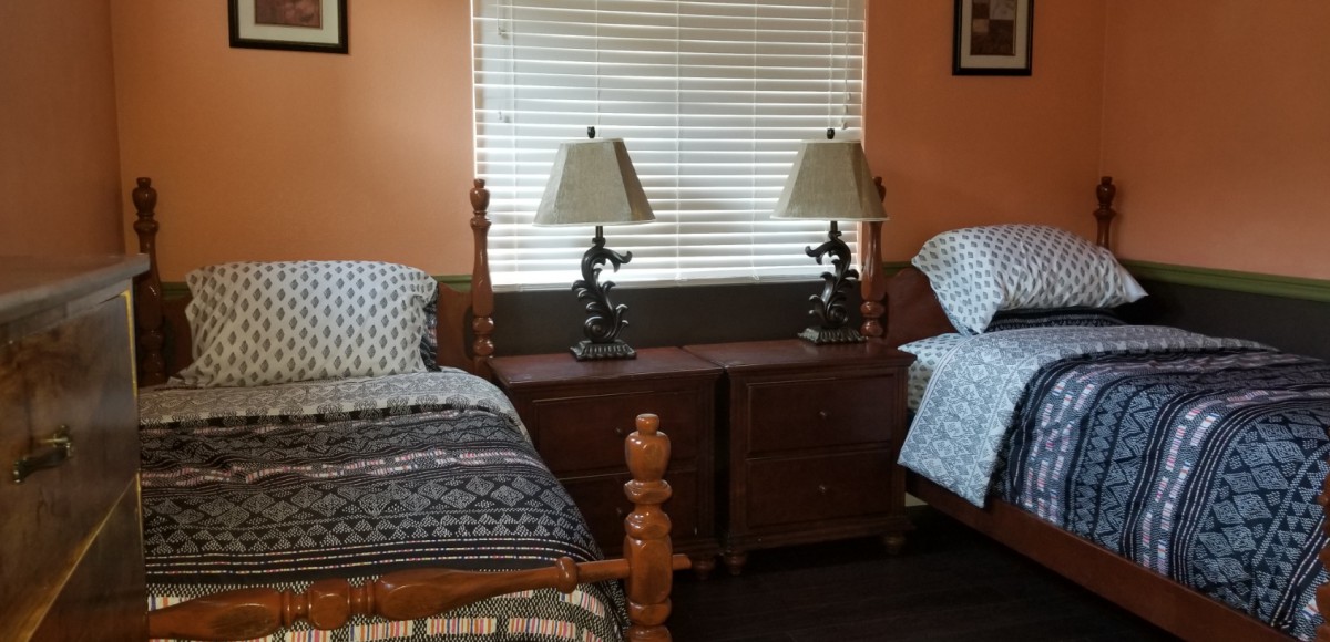 A cozy bedroom features twin beds with decorative lamps on a shared nightstand beneath a window with blinds.