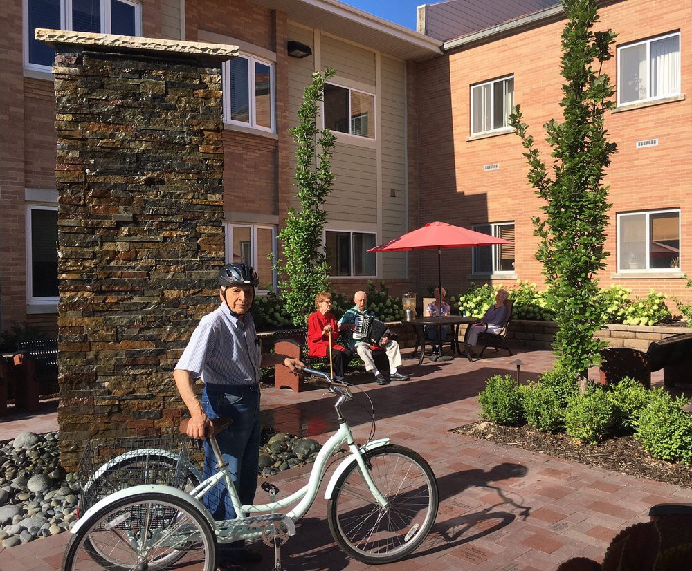 A man with a tricycle in a sunny courtyard area, with people sitting nearby.