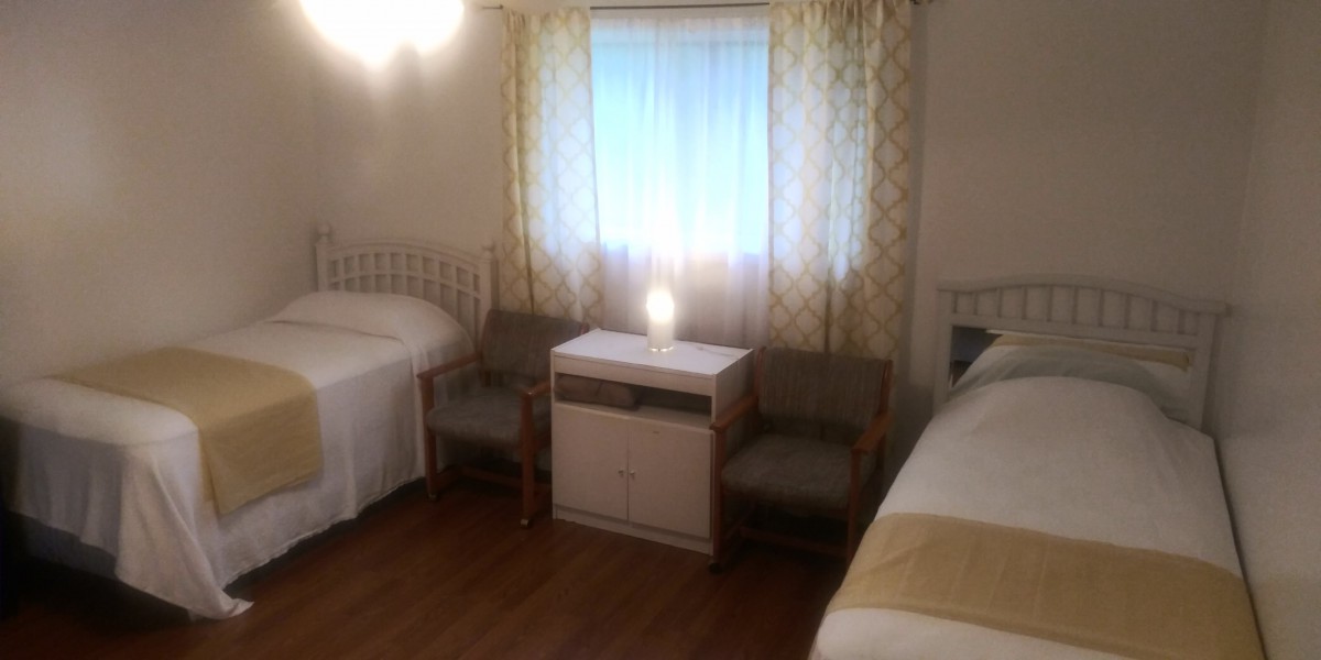 A cozy bedroom with two beds, separated by a nightstand with a lamp, and two chairs near a window with patterned curtains.