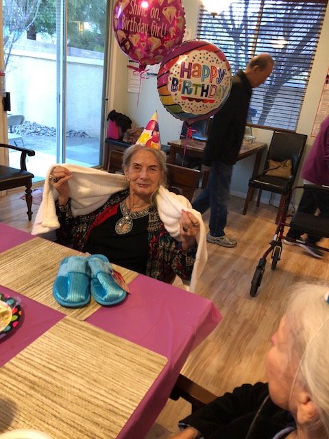 An elderly woman celebrating her birthday with a festive hat and balloons indoors.