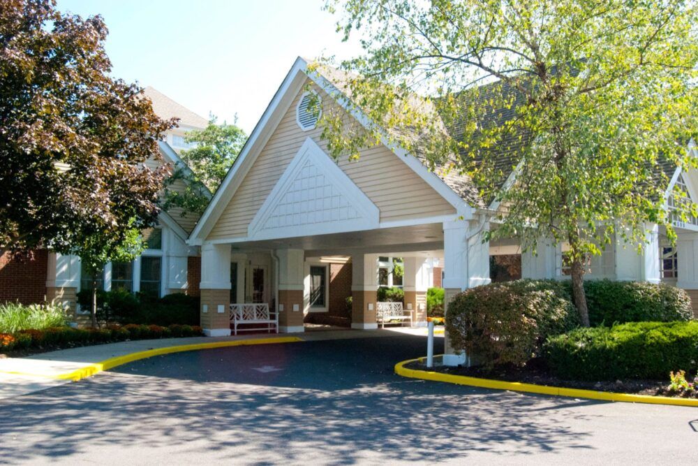 A building entrance with a sloped roof, surrounded by landscaped greenery and a paved driveway.