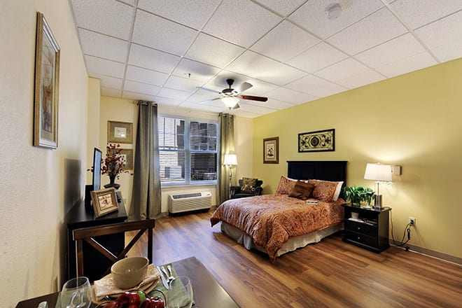A cozy bedroom with a wooden floor, ceiling fan, and natural light from a large window.