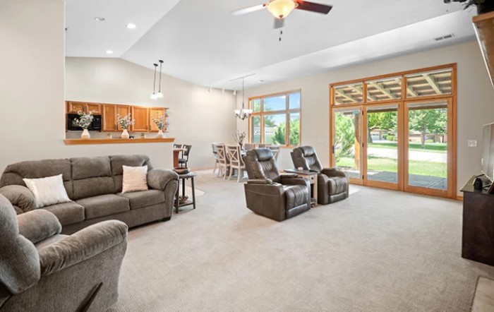 A spacious living room with modern decor, featuring sofas and recliners, a ceiling fan, and large windows allowing natural light.