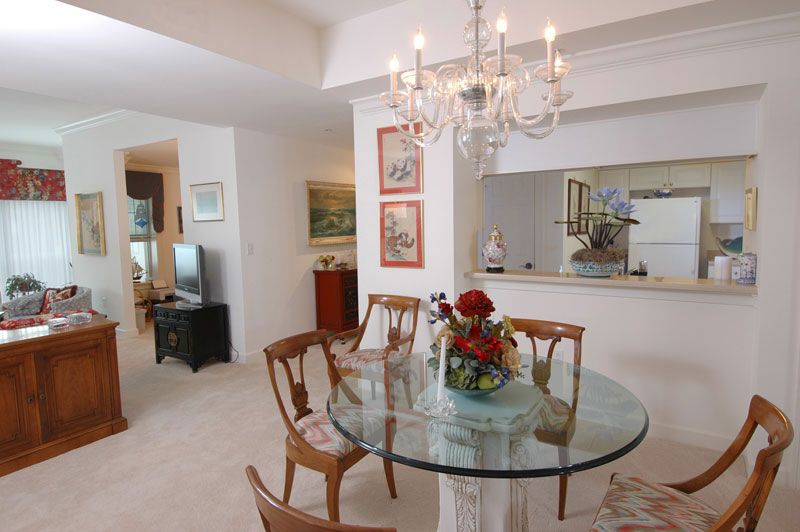A cozy dining room with a glass table and wooden chairs, illuminated by a chandelier and decorated with flowers.
