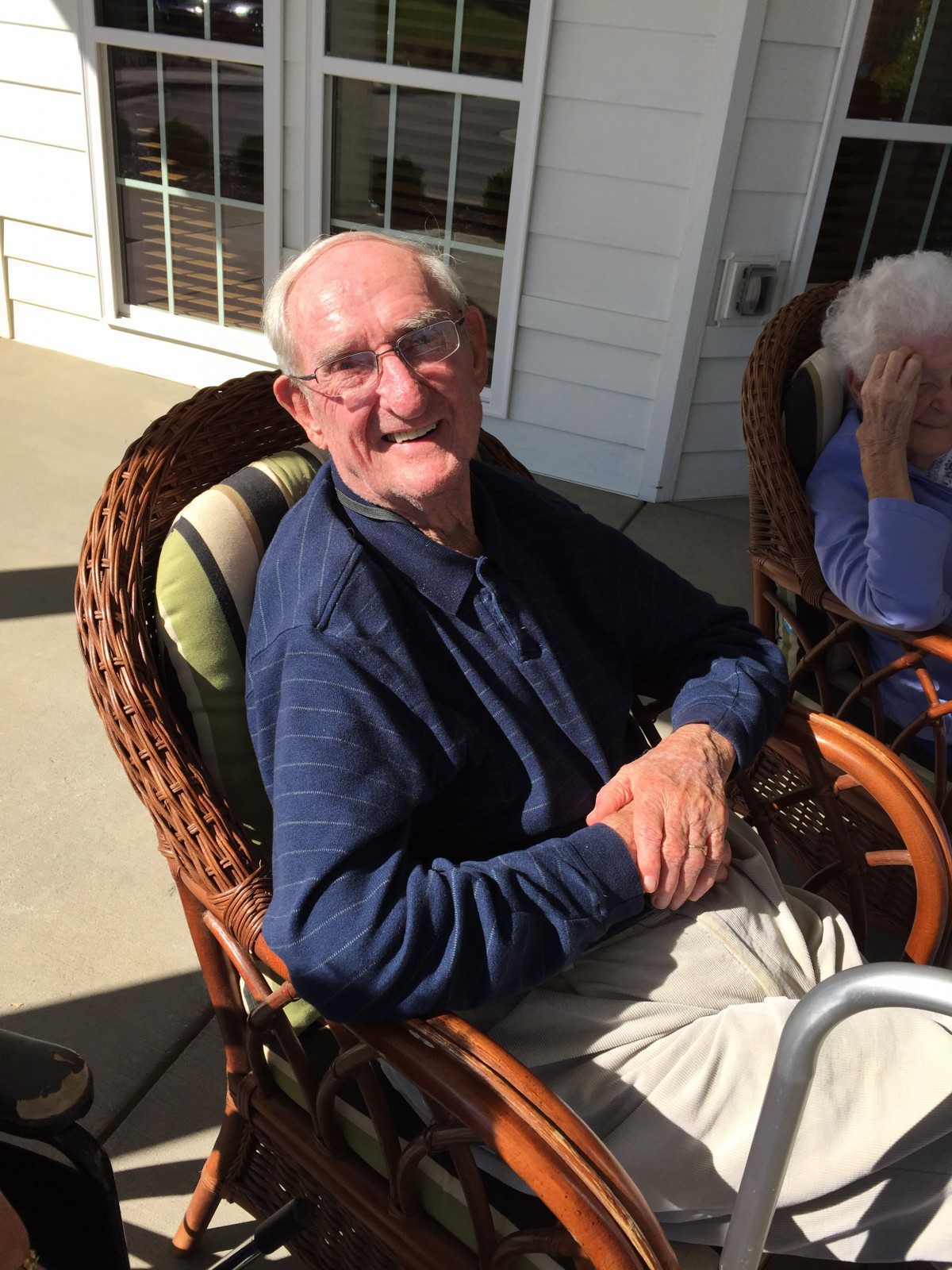 An elderly man is smiling while sitting outdoors on a sunny patio in a wicker chair.
