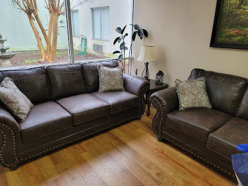 A cozy living room setup featuring a dark gray sofa and armchair, complemented by decorative pillows and natural light coming through the windows.