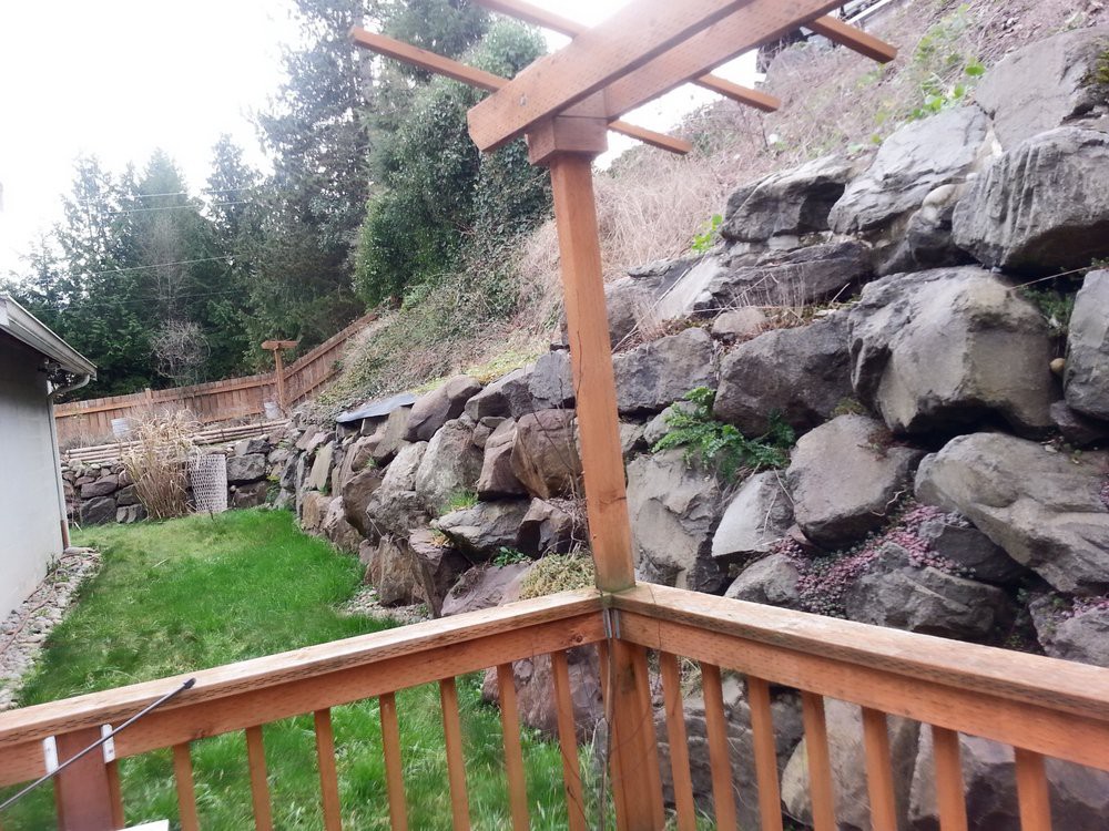 A backyard view with a wooden deck and fence overlooking a rock wall and trees.