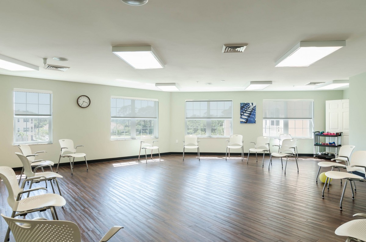 An empty room with chairs arranged along the walls, featuring large windows and a wooden floor.
